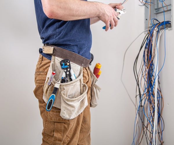 Electrician builder at work, examines the cable connection in the electrical line in the fuselage of an industrial switchboard. Professional in overalls with an electrician's tool.