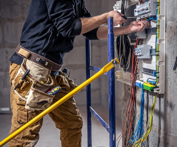 A male electrician works in a switchboard with an electrical connecting cable, connects the equipment with tools.