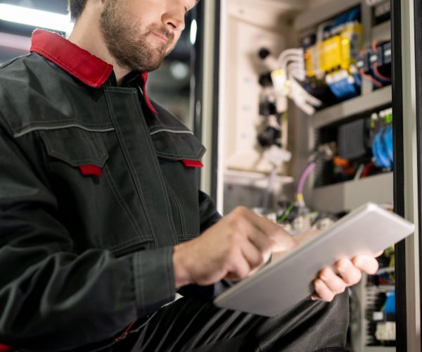 Young bearded engineer in workwear and hardhat checking settings of industrial machine while scrolling through technical data in tablet